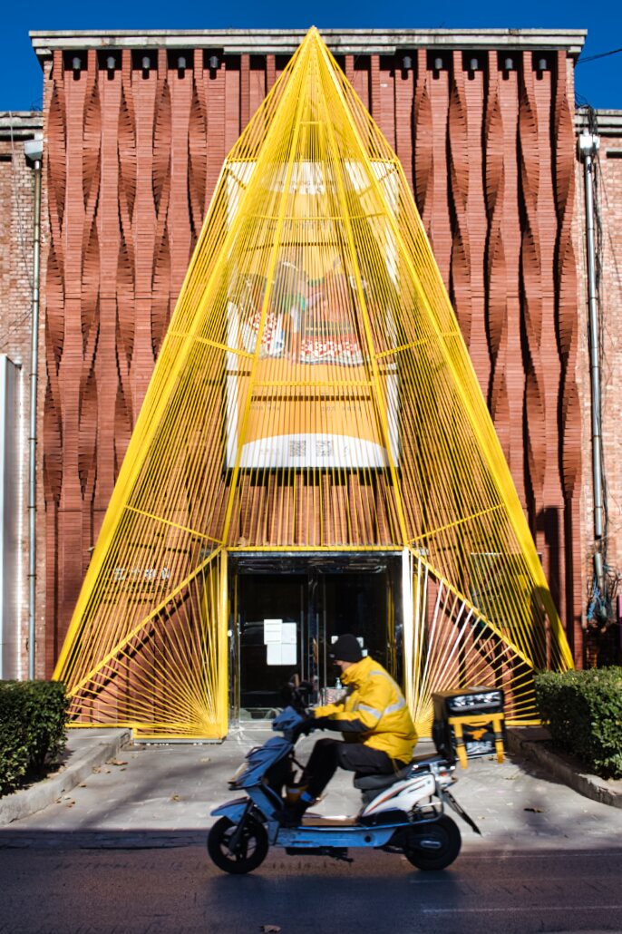 Scooterfahrer mit gelber Jacke vor einem Gebäude mit dreieckiger Draht/Metall-Deko im gleichen Gelb.