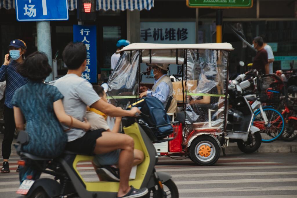 Das Farbfoto zeigt eine Pekinger Straßenszene an einem Zebrastreifen: ein Scooter, auf dem eine dreiköpfige Familie sitzt, ein Tuktuk, in dem ein betagtes Ehepaar fährt. Im Hintergrund Leihfahrräder und Fußgänger.